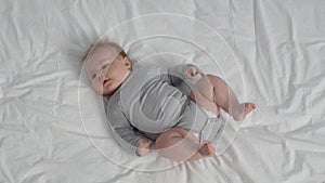 Cute Plump Newborn Baby In Grey Bodysuit Lying On Bed At Home
