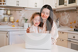 Cute portrait of mother and daughter sitting in a kitchen table and using computer