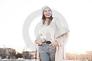 Cute positive young woman with beautiful smile in a fashionable knitted warm hat in a stylish milk-colored fur coat made of faux