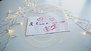 Cute postcard with declaration of love in calligraphic style rests on table with flickering garland.