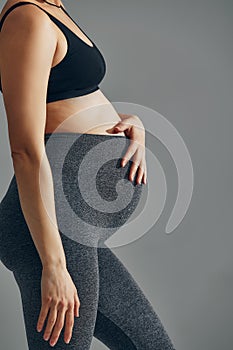 Cute pregnant belly isolated on gray. Side view of young pregnant woman embracing her abdomen with hands. Big belly on