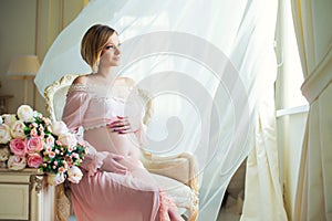 Cute pregnant woman sitting on chair near billowing curtains and hugs belly with love