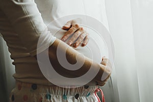 Cute pregnant woman in spotted pyjamas is stroking her belly near the window. Tender photo and happy pregnancy hug. Home