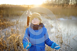 Cute preschool boy is playing on the ice of a frozen lake or river on a cold sunny winter sunset. Child having fun with icicle and