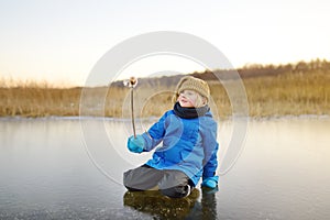 Cute preschool boy is playing on the ice of a frozen lake or river on a cold sunny winter sunset. Child having fun with icicle and