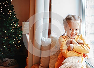 A cute preschool girl sitting on the windowsill with her plush toy waiting for Christmas near the christmas tree