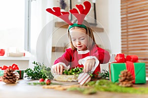 Cute preschooler girl dressed in reindeer costume wearing reindeer antlers making christmas wreath in living room.