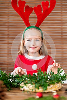 Cute preschooler girl dressed in reindeer costume wearing reindeer antlers making christmas wreath in living room. DIY Christmas.