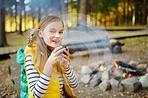 Cute preteen girl drinking tea and roasting marshmallows on stick at bonfire. Child having fun at camp fire. Camping with children