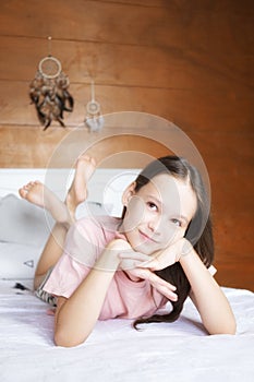 Cute preteen girl in pink t-shirt dreaming on white bed in boho style room against wooden wall and dreamcatchers. Scandinavian in