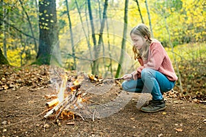Cute preteen girl roasting marshmallows on stick at bonfire. Child having fun at camp fire. Camping with children in fall forest