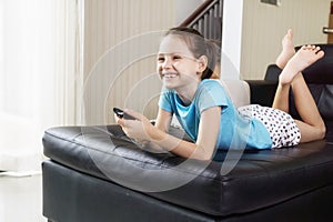 Cute preteen girl watching TV on couch using remote control. Living room interior in background