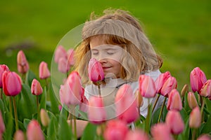 Cute pretty blonde kid with eyes closed smelling tulip flower in the Spring park. Outdoor portrait of cute blonde kid