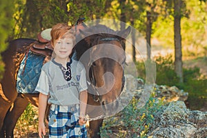 Cute red hair boy smiling to camera and standing in forest with horse