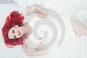 Cute red hair woman in cozy knitted white sweater dress relax on the fur bed in her white room