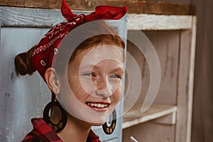 Cute red-haired girl with freckles, dressed in a red bandana and a plaid shirt on the background of the kitchen. Advertising, pin-