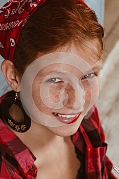 Cute red-haired girl with freckles, dressed in a red bandana and a plaid shirt on the background of the kitchen. Advertising, pin-