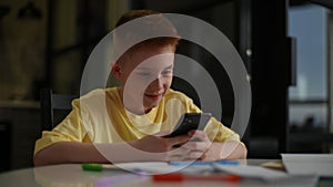 Cute redhead child boy with freckles in yellow t-shirt sitting at table holding smartphone enjoying using mobile apps