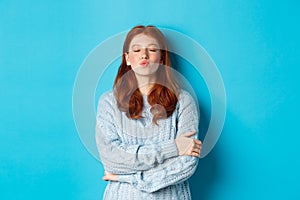 Cute redhead teen girl waiting for kiss, pucker lips and close eyes, standing in sweater against blue background