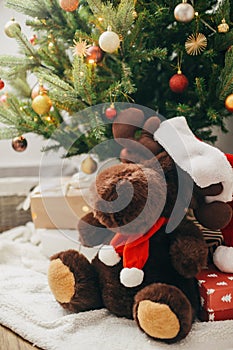 Cute reindeer toy in santa hat under modern decorated Christmas tree with gifts and festive lights. Big surprise toy at christmas