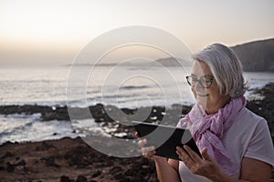 Cute relaxed senior woman gray hair sitting near the beach surfing the net with tablet. Dusk light. Mountains and sea in