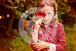 Cute sad child girl hiding behind red autumn leaf in the garden