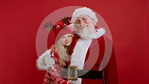 Cute Santa Claus in Red Outfit Hugs Little Blonde Girl in Red Cap and Sweater, Smiling and Waving Their Hands on Red