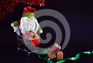 Cute Santa Claus toy, sitting in red shopping cart. Christmas sale or New Year concept, isolated on black background