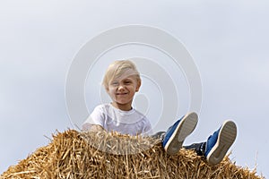 Cute schoolboy boy is sitting on haystack in field on sky background. Harvesting. Summer day in village