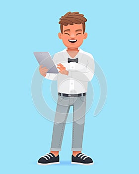 A cute schoolboy child in a white shirt with a bow tie is standing with a tablet in his hands. Happy little boy