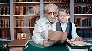 Cute schooler boy and 70s grandfather reading letter together doing homework at public retro library