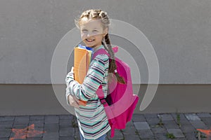 A cute schoolgirl with a knapsack behind her back smiles and holds colorful notebooks and folders in front of the school. Children