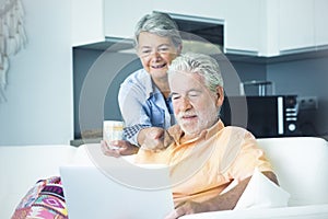 Cute senior man and woman using computer at home. Man sitting on the sofa and woman behind him watching the laptop display. Mature