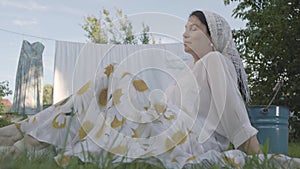 Cute senior woman with a white shawl on her head resting in the garden sitting on the grass in front of the clothesline