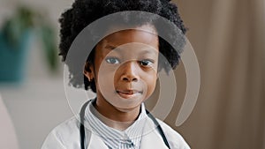 Cute serious african american kid girl in medical clothes dressed in white coat standing indoors posing looking at