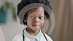 Cute serious african american kid girl in medical clothes dressed in white coat standing indoors posing looking at