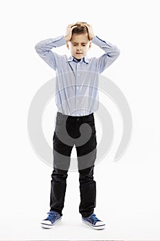 A cute serious boy, an 8-year-old schoolboy in a blue shirt and trousers, is holding his head in his hands. Full height. White