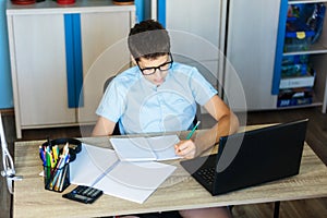 Cute serious boy in blue shirt sitting behind desk in his room next to laptop and study. Teenager in earphones makes homework,