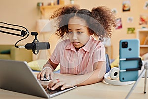 Cute serious schoolgirl looking at laptop screen while recording new podcast