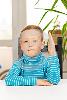 Cute seven-year-old child boy sits at home at a white wooden table against the background of a light window. Selective focus. Clos