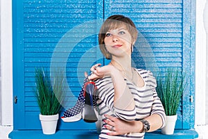 Cute short-haired teen Girl holding her new high heeled shoes in front of blue jalousie window with green plants. Shopping concept