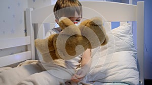 Cute sick boy in protective medical mask lying in bed with teddy bear. Concept of child virus and kids protection during