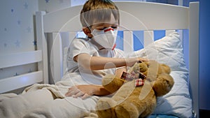 Cute sick boy in protective medical mask lying in bed with teddy bear. Concept of child virus and kids protection during