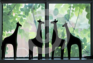 Cute silhouettes of giraffe figures standing on a window ledge with blurred greenery and flowers showing through