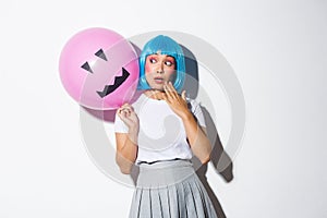 Cute and silly asian girl in blue wig, celebrating halloween, looking surprised at balloon with scary face