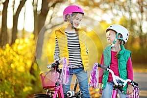 Cute sisters riding bikes in a city park on sunny autumn day. Active family leisure with kids. Children wearing safety hemet while