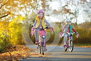 Cute sisters riding bikes in a city park on sunny autumn day. Active family leisure with kids. Children wearing safety hemet while