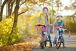 Cute sisters riding bikes in a city park on sunny autumn day. Active family leisure with kids. Children wearing safety hemet while