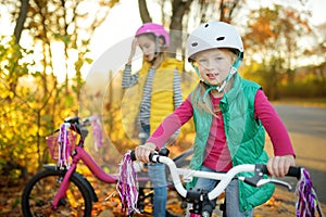 Cute sisters riding bikes in a city park on sunny autumn day. Active family leisure with kids. Children wearing safety hemet while