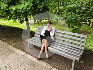 Cute slender woman in a business suit sits on a bench in the park with a laptop and speaks by phone. An adult girl in glasses